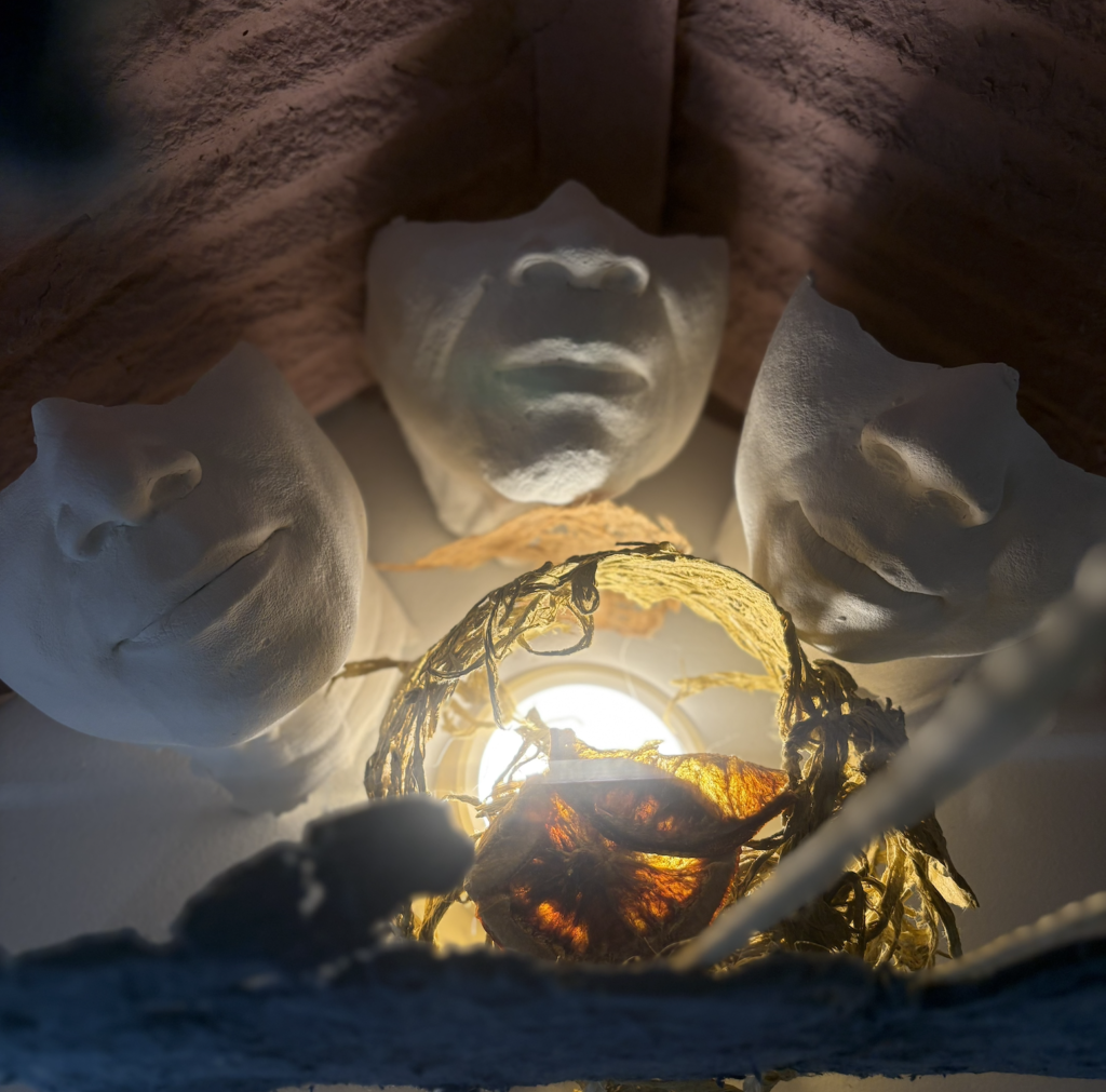 Close-up of three white sculpted face masks arranged in a triangular formation under a warm light. In the foreground, a delicate structure made of dried plant material and translucent fibers surrounds the light source, casting soft shadows on the masks.